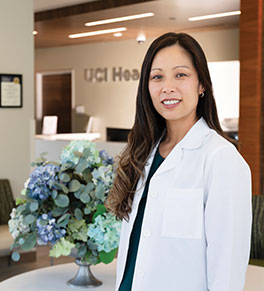UCI Health menopause expert Dr. Rebecca Sauer wears a white coat and stands near a floral arrangement at a UCI Health clinical office.