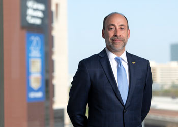 uci health ceo chad lefteris wearing dark suit, blue tie, white shirt, standing in front of window with uci medical center in the background