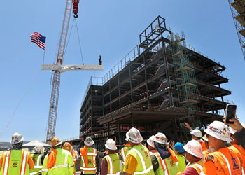construction workers, uci health co-workers and leadership wearing  hard hats and vests watch as the final beam is put in place at uci health irvine, topped with a tree and an american flag