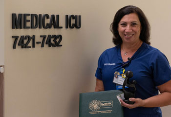 uci health nurse and daisy award winner janette rhodes sanches in front of medical icu sign wearing scrubs and badge, holding award