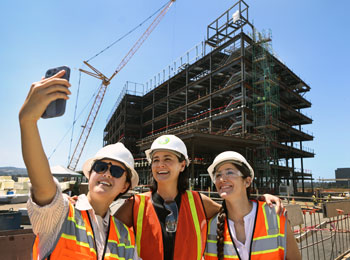 uci health co-workers wearing orange construction vests and hard hats stand in front of the under construction uci health irvine on a sunny day