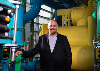 uci health director of facilities joe brothman wearing a dark suit, white shirt standing in the central utilities plant in front of blue and yellow pipes