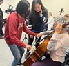 uci student wearing red sweatshirt and uci student wearing blue sweatshirt helping senior woman play a stringed instrument