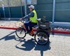 a man wearing a white hat, dark shirt and pants and a yellow construction safety vest rides an electric bike on a paved road in front of a red curb and fence