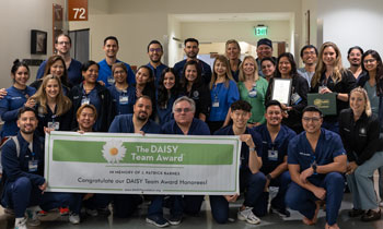 the uci health uci medical center cardiovascular intensive care unit team gathers in scrubs for a photo in a hallway holding a sign saying the daisy team award