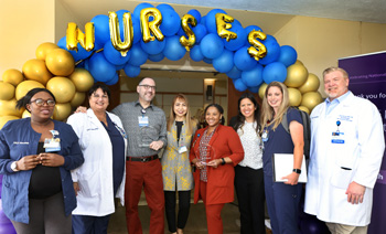 a group of nursing excellence award attendees stand under yellow and blue balloons with gold balloon letters spelling out nurses