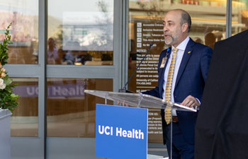 uci health president and ceo chad t lefteris stands behind a lectern outdoors with a uci health sign speaking into a microphone while wearing a dark suit and yellow tie