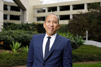 uci health chief financial officer randolph siwabessy wearing a dark suit and tie standing outside in front of a structure and a green lawn and plants