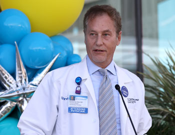 uci health cancer center director dr richard van etten stands and speaks at a microphone in front of blue and yellow and silver balloons wearing a white coat, tie blue shit and name tag