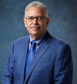 uci health chao family comprehensive cancer center director miguel villalona calero in front of a blue background wearing a blue sport coat and blue tie
