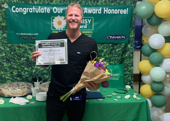 daisy award winner uci health los alamitos nurse james needham holding his certificate and a bouquet of flowers standing in front of balloons and a sign saying congratulations to our daisy award honoree
