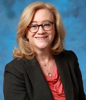 ramona agrela wearing a dark jacket and red top in front of a blue background; she has been named one of becker's chros to know
