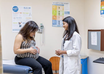 a physician in a white coat smiles at a pregant patient who is looking at her belly during an appointment at uci heath family health center anaheim