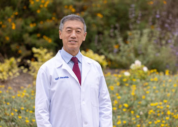 uci health integrative oncologist dr gary deng poses in his white coat in front of green bushes