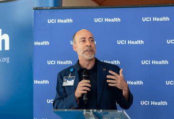 uci health president and ceo chad lefteris speaks to a crowd at an event in front of a blue and white uci health step and repeat