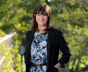 Huntington's disease researcher Leslie Thompson, wearing a black sweater and a blue patterned blouse, stands in a tree-lined area of UC Irvine.