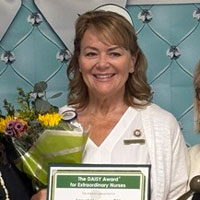 uci health nurse janet hewson wears a nurse's white cap as she holds her daisy award for nursing excellence and a bouquet of flowers