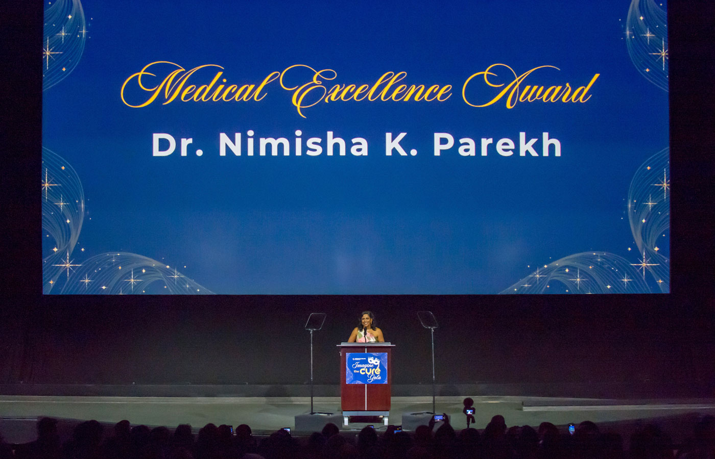 uci health gastroenterologist dr. nimisha parekh stands on stage in front of a large screen that says medical excellence award dr nimisha parekh, during a crohn's and colitis foundation gala