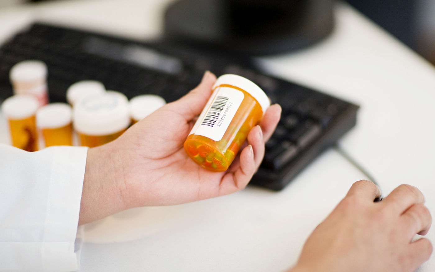 Hand with white-coat sleeve holds a medication bottle while using a computer mouse
