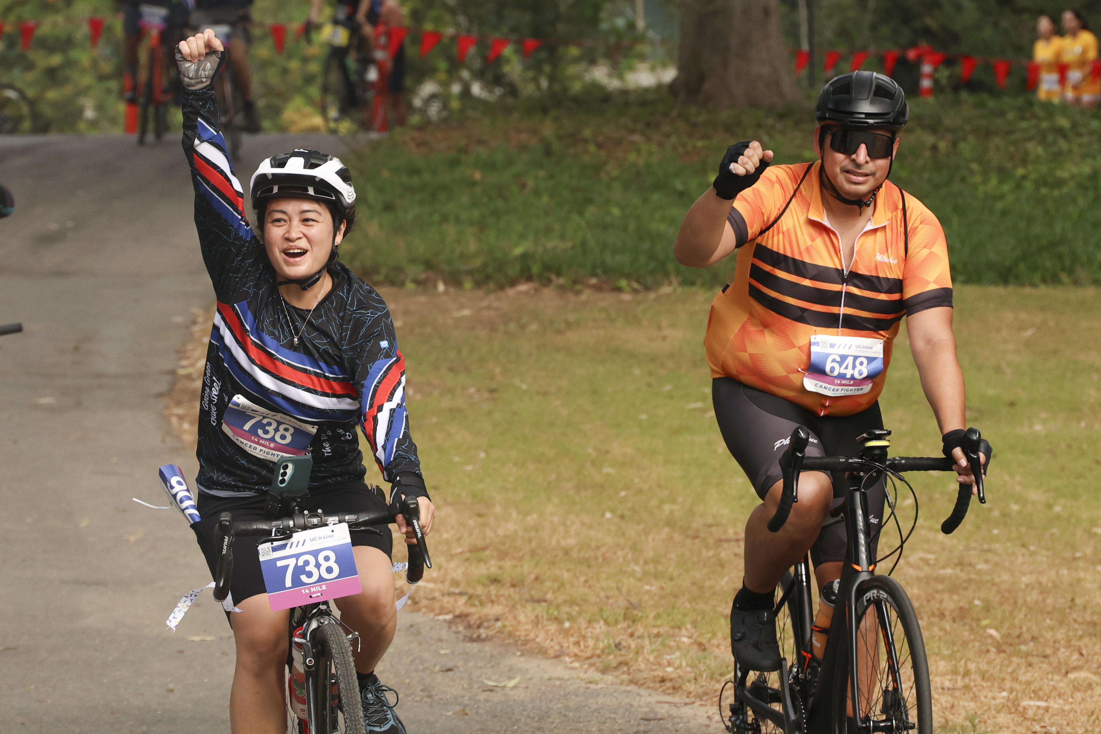 two cyclists pump their fists as they ride on a paved path during the 2026 uc irvine anti-cancer challenge 