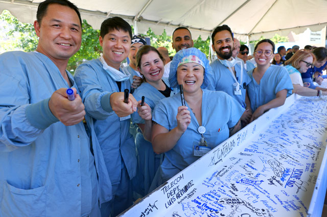 seven uci health co-workers hold up their pens and smile after signing a white beam covered in signatures before the beam tops off uci health irvine