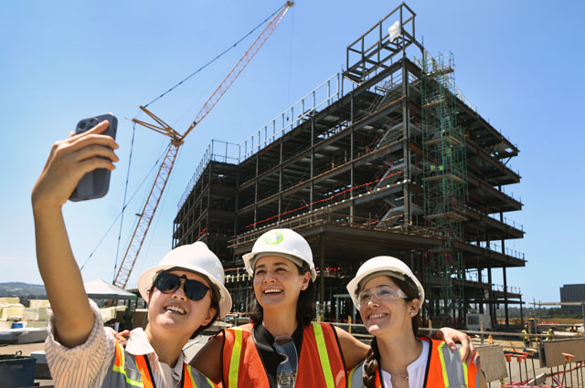 uci health - irvine construction topping off ceremony; uci health coworkers wearing vests and hard hats take a selfie in front of the final building under construction