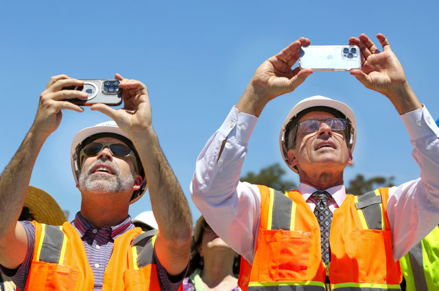 uci health - irvine construction topping off ceremony; uci health ceo chad t lefteris and uci school of medicine dean dr michael stamos wearing orange vests and hard hats take a photo of the final beam being lifted into place