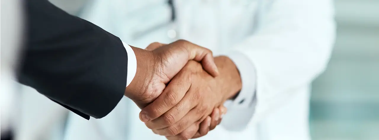 Two people shaking hands; one in a black suit, the other in a white coat, on a blurred background.