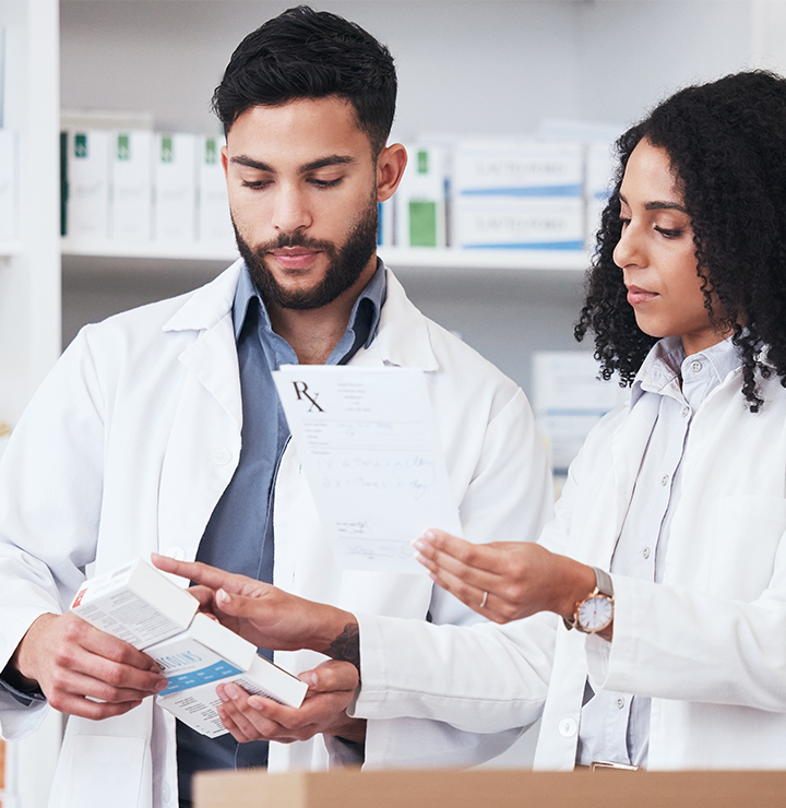 Two pharmacists behind a counter review a prescription with shelves of medication boxes in the background.