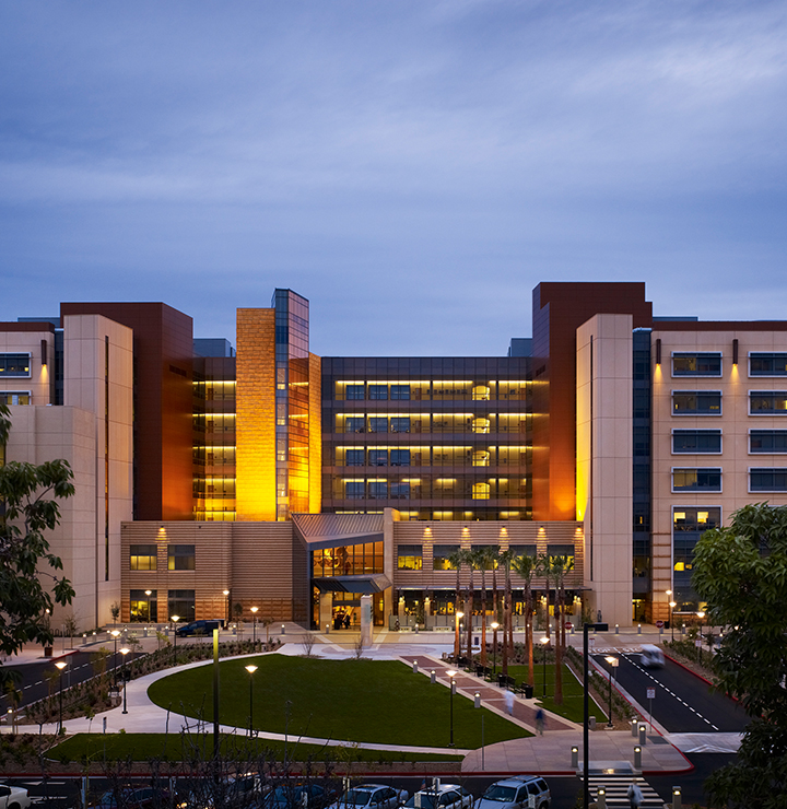 Modern hospital building at dusk, illuminated with warm lights, surrounded by trees.