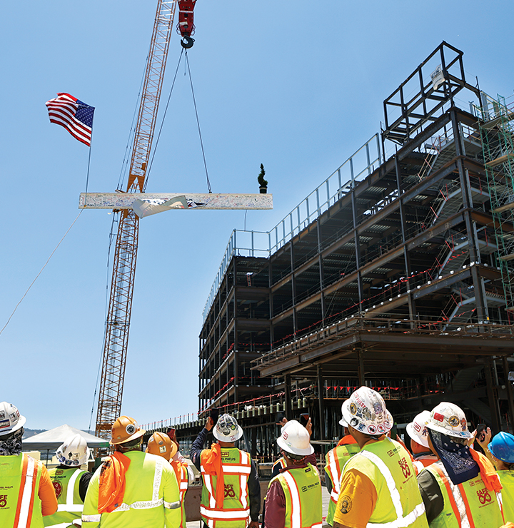 Construction workers in hard hats and vests observe a multi-story building under construction with a crane, under a clear blue sky.