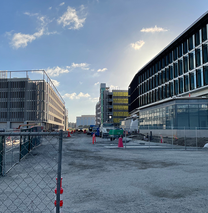 Construction site with modern buildings under a partly cloudy sky, and machinery along the fenced pathway.