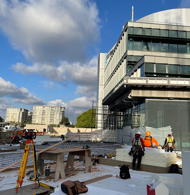 Construction site with workers wearing safety gear, handling materials and equipment. Modern building and residential towers in the background.