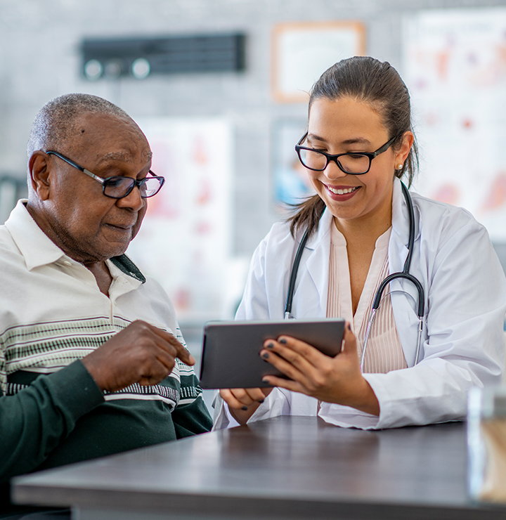 Doctor and elderly patient discussing information on a tablet in a medical office, both smiling and wearing glasses.