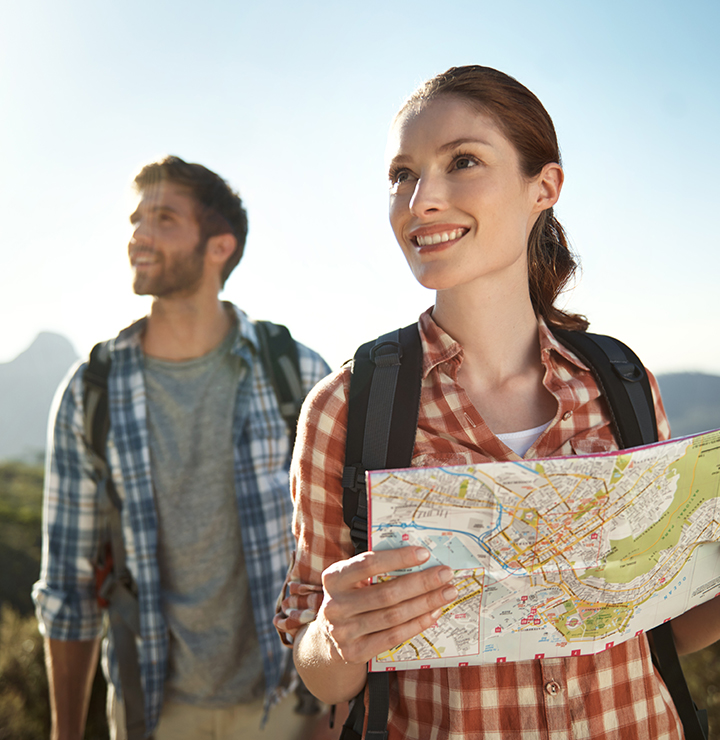 A smiling woman holding a map and a man with a backpack are hiking in a scenic mountainous area under a clear sky.