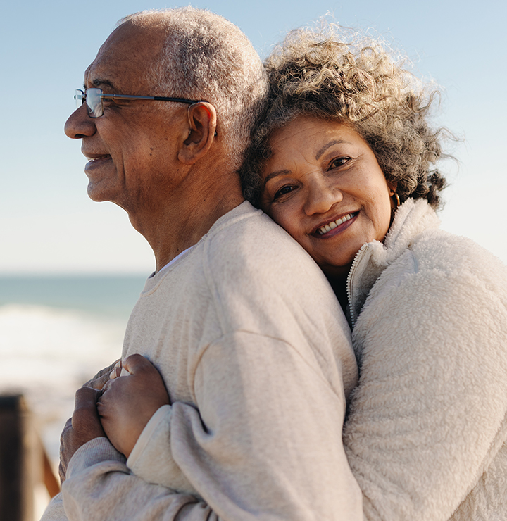An elderly couple embraces while standing on the beach, smiling and looking content with the ocean in the background.