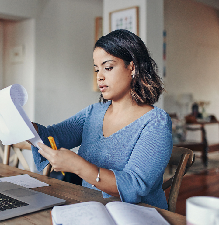A woman in a blue top sits at a table, holding papers and a pen, with a laptop in front of her.