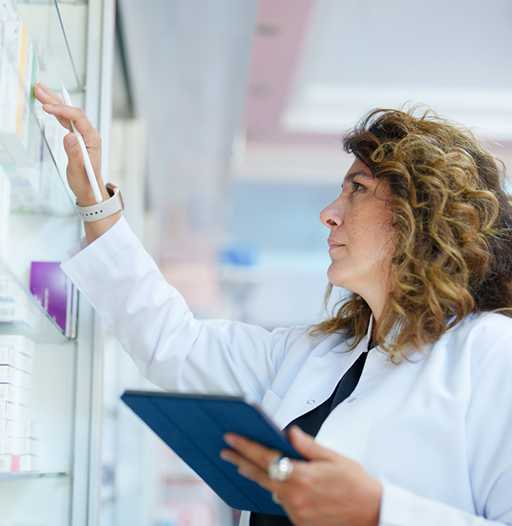 A woman in a white lab coat examines items on a shelf while holding a tablet.