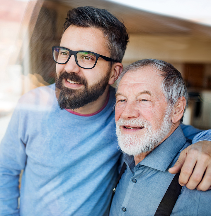 Older man with gray hair and beard smiles while being embraced by a younger man with glasses, both looking contently out of a window.