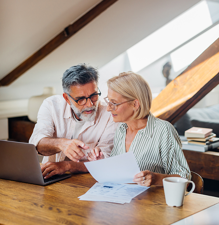 Older couple reviewing documents together at a wooden table, with a laptop and a mug nearby, under slanted attic ceilings.
