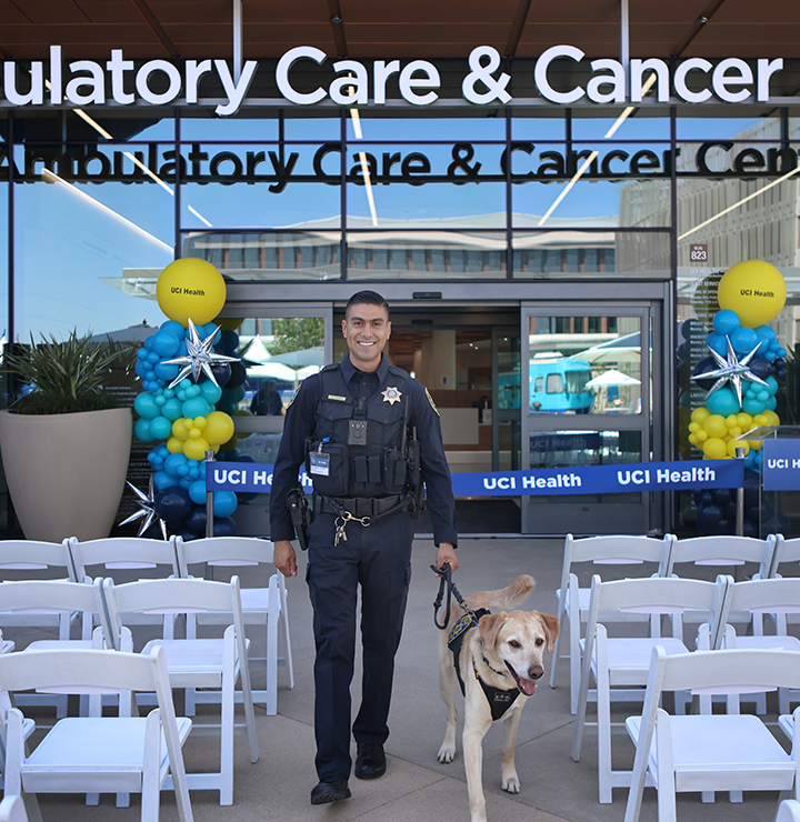 Police officer with a dog stands in front of a UCI Health building decorated with balloons and ribbon, facing rows of empty chairs.