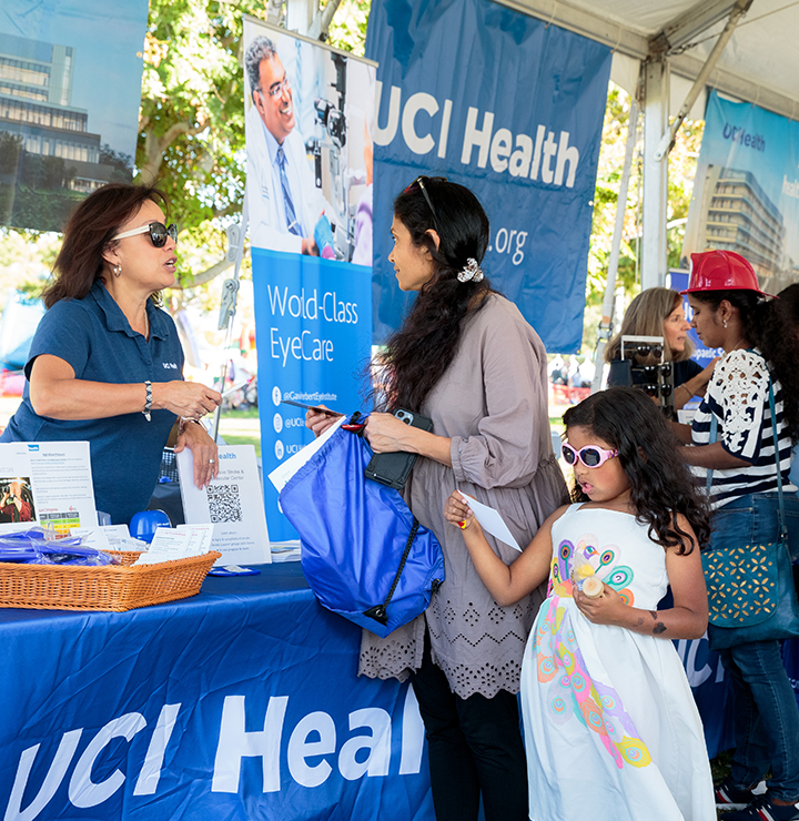 Two women talking at a health fair booth with informational banners, one wearing a blue shirt and the other holding a blue and white bag.