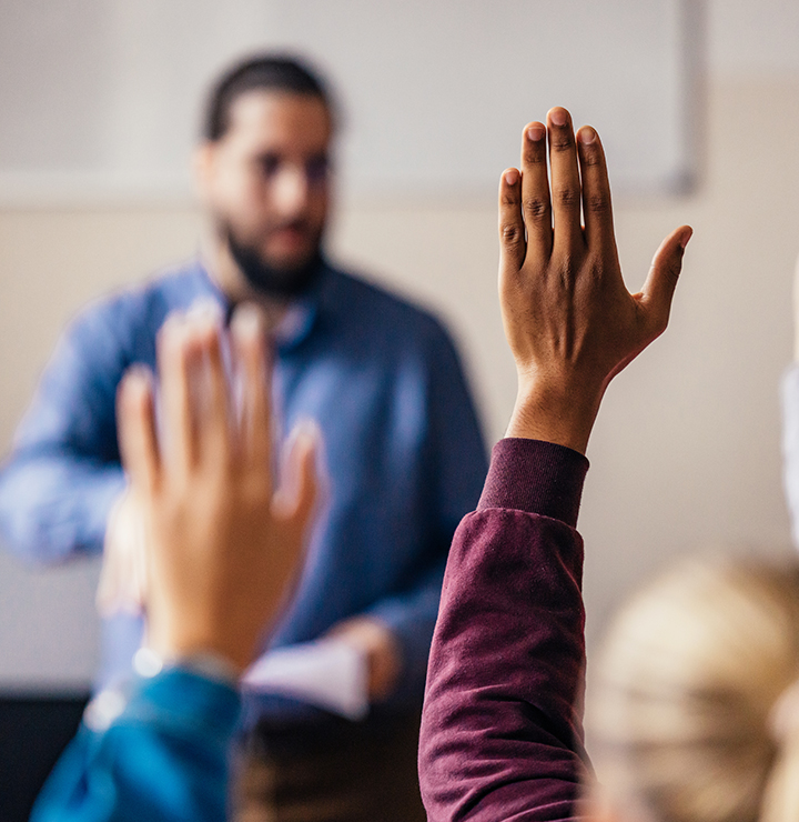 Students raise hands in a classroom while a teacher stands in the background, slightly out of focus.