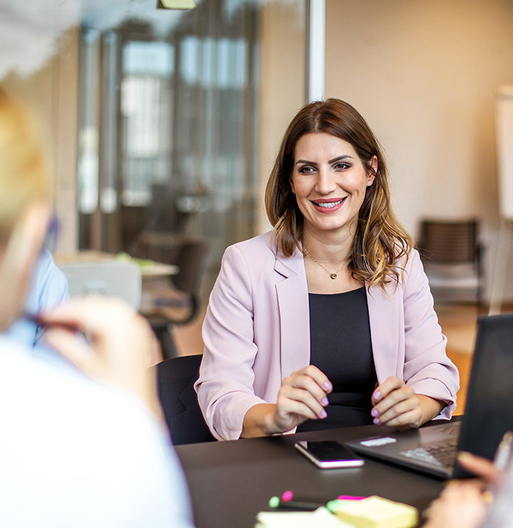 A smiling woman wearing a pink blazer sits at a table in an office setting, engaged in conversation with colleagues.