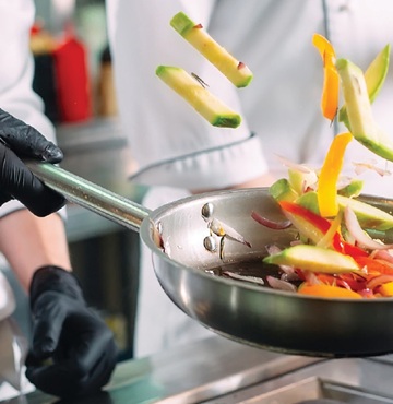 A black-gloved hand flips a skillet of vegetables