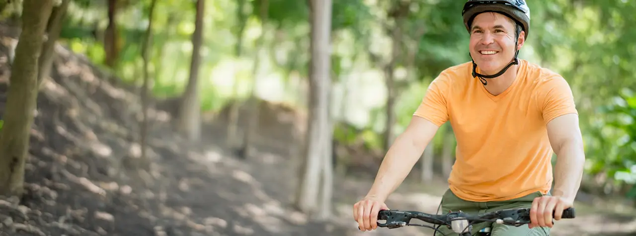 Smiling man wearing a helmet rides a bicycle on a forest trail, surrounded by lush green trees.
