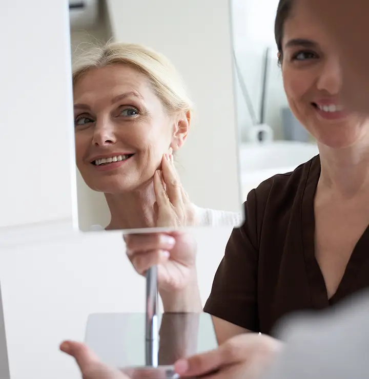 Blonde woman smiles, touching her face while looking in a mirror with another woman beside her.