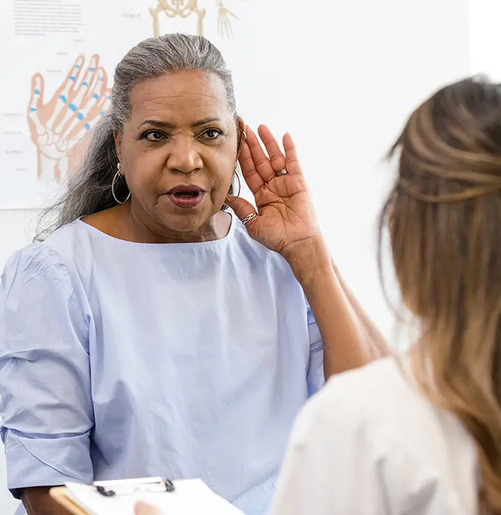 Woman with gray hair gestures to her ear while speaking in a medical office.