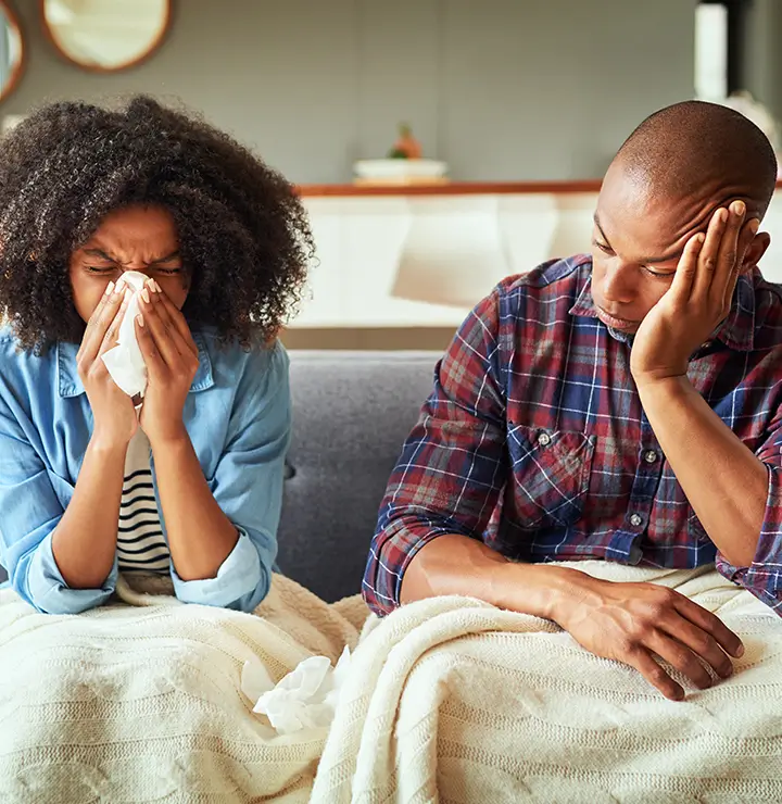 Woman sneezes into a tissue on a couch while a man rests nearby under a blanket.