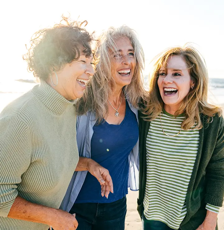 Three women laugh together on a sunny beach with the ocean in the background.